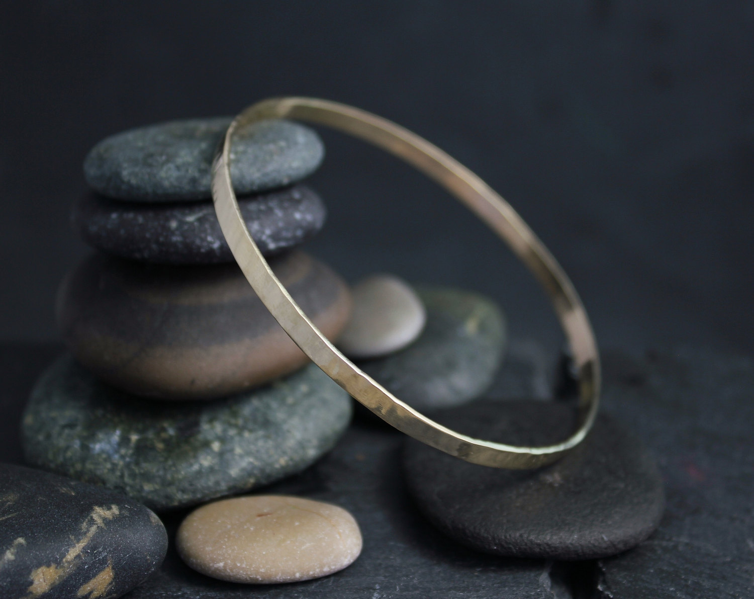 Hammered 14k yellow gold bangle bracelet resting against stacked river stones
